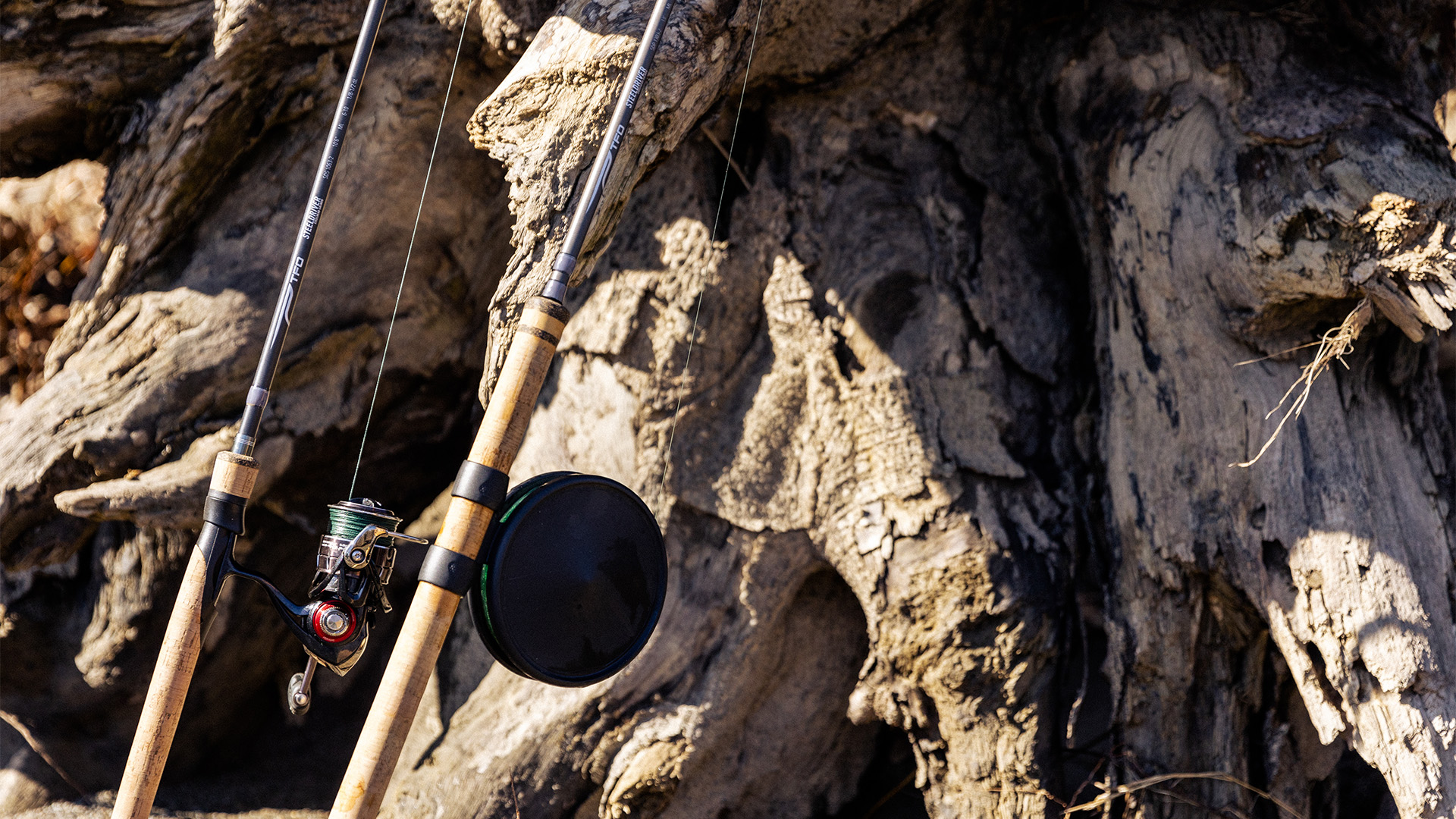 Close-up of TFO Steeldriver spinning and Centerpin rods resting against a large driftwood log, showing handle detail and reel setup for fall salmon and steelhead fishing.
