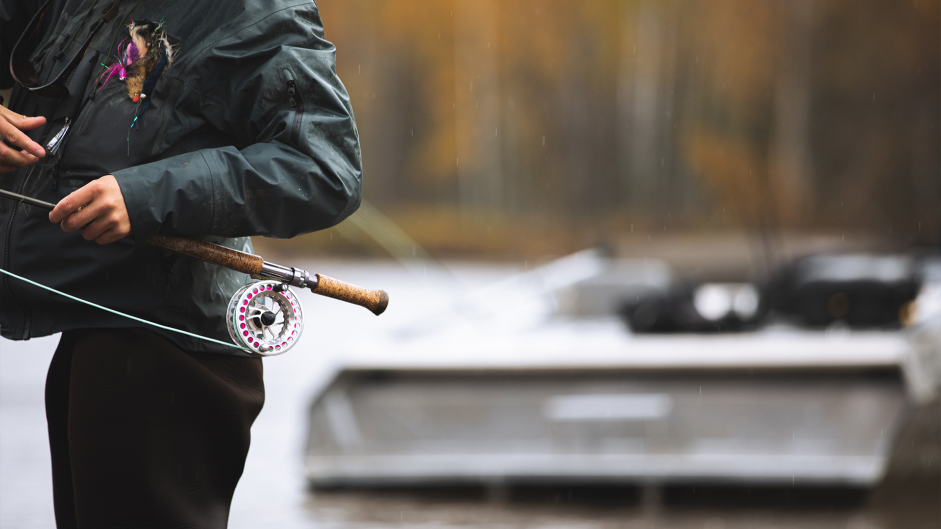 Close-up of an angler holding a TFO LK Legacy Two Handed fly rod with bright pink fly line and streamer patterns attached to a jacket, with a drift boat blurred in the background.