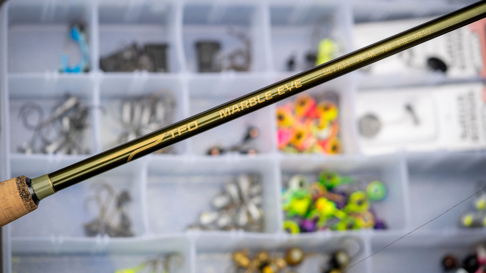 Close-up of a TFO MarbleEye fishing rod resting over a tackle box filled with colorful jigs and weights.