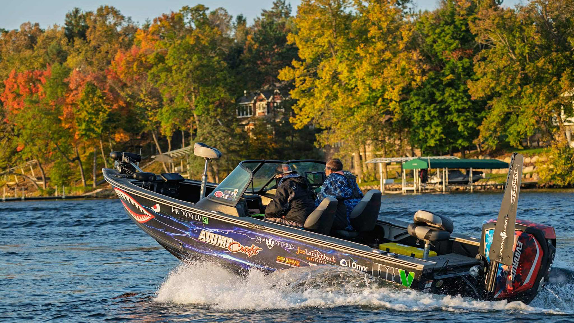Two anglers motor across a lake in a tournament boat during fall, surrounded by colorful shoreline trees and crisp morning light.