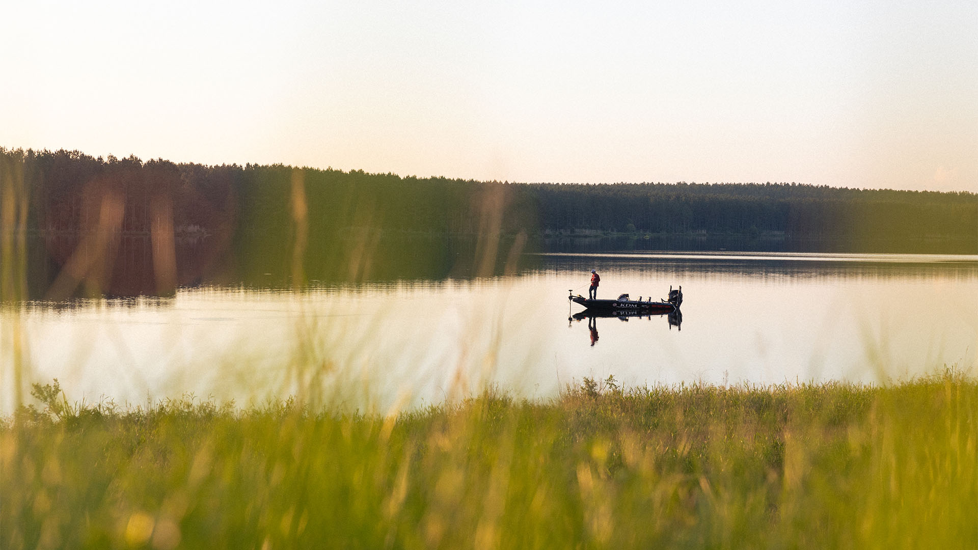 Bass boat at sunrise on a calm lake with an angler casting toward the shoreline.