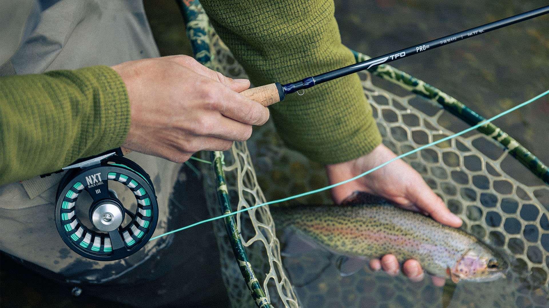 Close up of an angler landing a rainbow trout with a TFO Pro III fly rod and NXT Black Label reel, showing the rod’s grip and branding over a netted fish in a clear stream.